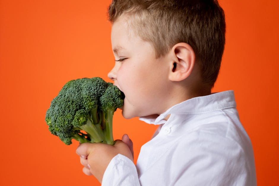 A young child biting into fresh broccoli against an orange background